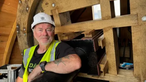 Andrew Turner/BBC Millwright James Forsyth is wearing a black tee shirt, a black wristwatch, a high visibility waistcoat and a white hard hat. He has a close cropped goatee beard, and is sitting in the cap of the wind pump next to the oak brake wheel, his arm rested on the iron drive shaft.