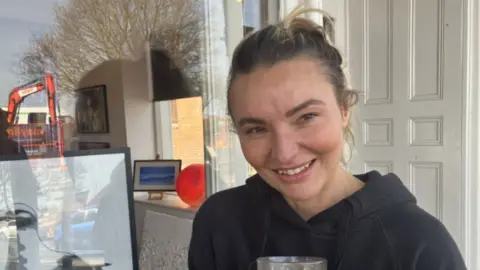 Lisa-Marie Robinson - a woman with dark blonde hair in a pony tail who is smiling. She is standing next to her art gallery window and there is a picture of a seagull in the window.