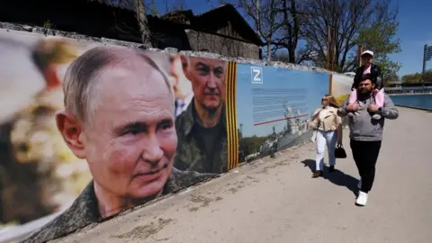 A poster showing Russian President on the side of a road where a man wearing black trousers, a gret top is carrying a child on his neck, with a woman walking by his side, wearing light blue trousers,a white top and a pink jacket, as well as carrying a small black handbag 