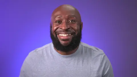 Riley wears a grey shirt and smiles at the camera in front of a blue background.