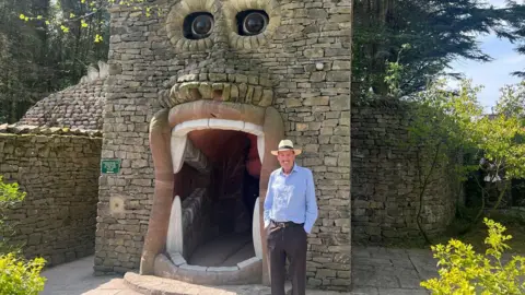 Georgia Levy-Collins/BBC An elderly man in a blue shirt and cowboy-style hat. He is smiling widely and has a moustache. He stands in front of a stone doorway in the shape of a giant mouth with eyes above and big teeth.