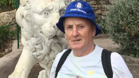 A picture of Stanley Smith. He is smiling and is wearing a blue bucket hat with the words Sergio Tacchini written on in white lettering. He appears as though he is on holiday in a park. A stone lion is behind him. He has a small stud earring and wears a white T-shirt. The straps of a black backpack can also be seen.