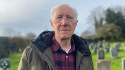 A man wearing a red and navy tartan shirt with a khaki green coat. He's standing in front of a number of gravestones.