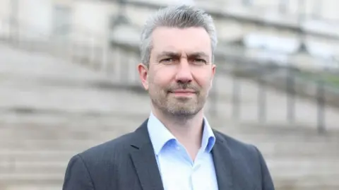 Alliance Party A man with grey hair and stubble, looking directly into the camera. He is waring a light blue shirt with the top button open and dark grey jacket