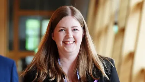 PA Media Gillian Mackay has long brown hair and wears a black blazer and lanyard