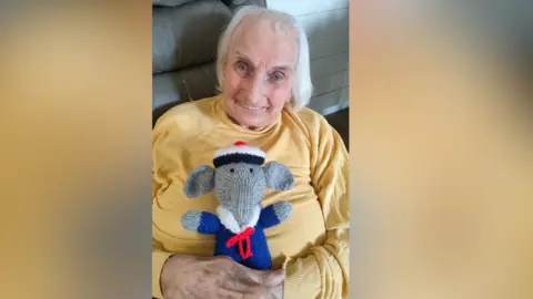 Hill Care Group Joyce Baxtrem has white hair and a yellow jumper. She is sitting on a grey chair holding a knitted, stuffed toy elephant wearing a sailor's outfit.