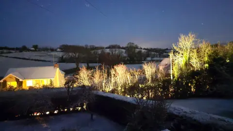 Colm - BBC weather watcher A house is lit up with yellow light as snow covers it, the fields and trees. The sky is a dark blue.