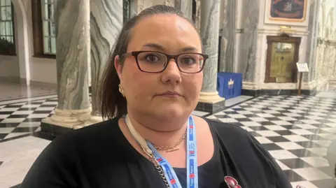 Sarah is standing in large room and in the background there is a black and white tiled floor. Sarah is wearing glasses and has her hair tied back 