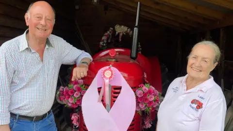 John Chapman is smiling at the camera and leaning on a red tractor decorated with pink ribbons and flowers in a barn. He is wearing a checked shirt and jeans. Annie Chapman is standing the other side of the tractor and is smiling. She is wearing a light pink polo shirt that says Ladies' Road Run.