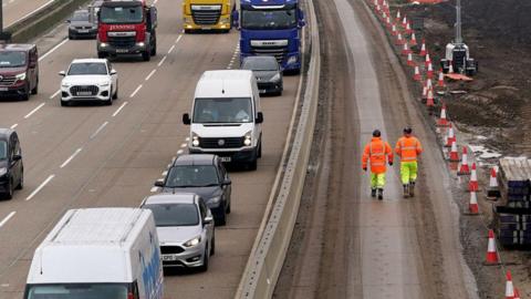Two workers in high vis jackets and hard hats walk between traffic cones and a busy motorway of queuing traffic  