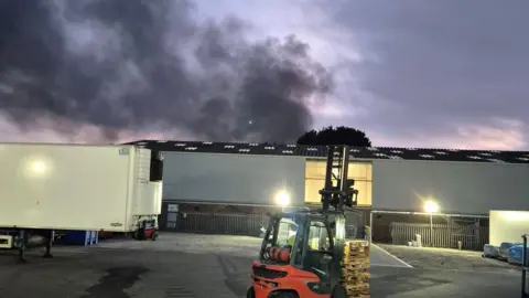 An industrial site with a large plume of black smoke in the distance. The back of a white lorry is to the left of the image.
