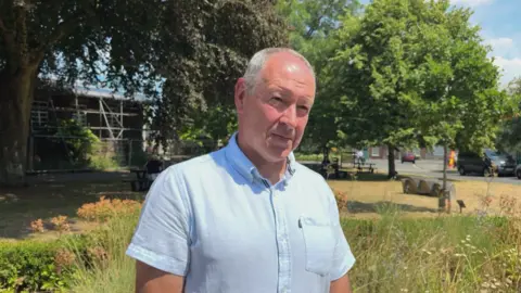 Nick is standing on the grass in front of Dorset Council HQ in Dorchester. He is wearing a blue short sleeve shirt