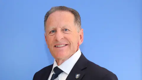 BRUFC A head an shoulders image of Phil Montanaro - he is wearing a dark suit and tie and a white shirt, he has short grey hair and is clean shaven