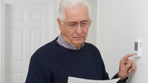 an older man with grey hair and wearing glasses, a blue jumper and checked shirt is reading an electricity bill and adjusting his thermostat