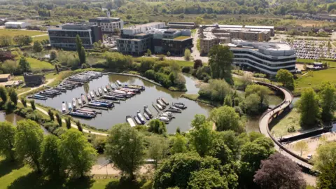 UON An aerial shot of the University of Northampton, showing several buildings, trees, water, narrow boats on the water, green areas, and cars parked to the right.