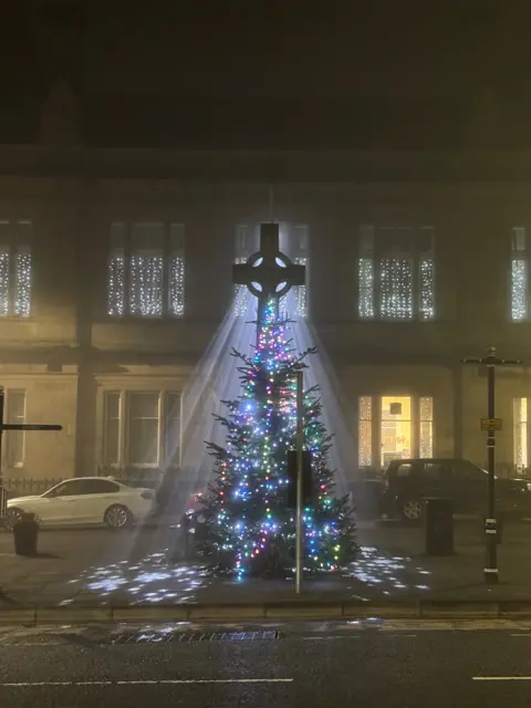 Amy Welsh White spotlight shining from a building onto a decorated Christmas tree in a town square.
