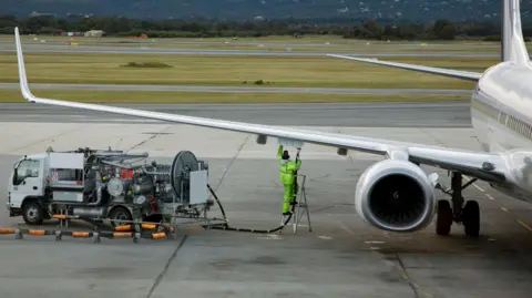 A man in a fluro yellow suit refuels an airplane sitting on an airport tarmac.