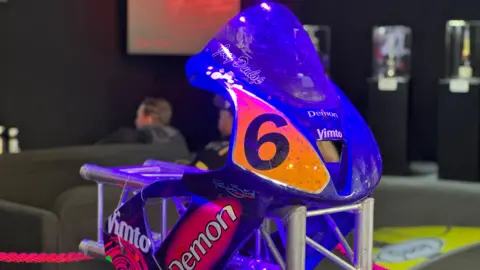 The blue bodywork of a motorcycle sits on a silver frame. It features a black number 6 on an orange background.