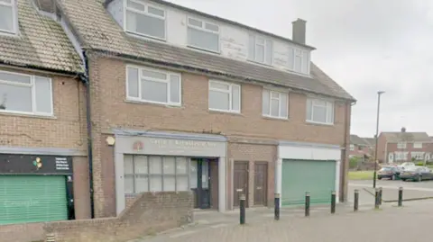 Google A two storey brown brick building with white dormer windows in the roof. On the ground floor, one half of the building has an office and the other a large green shutter across it. 

