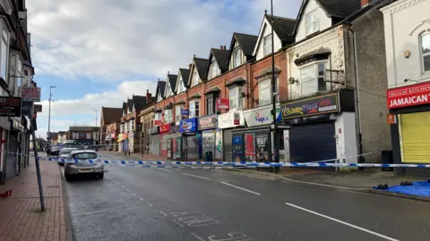 A police cordon stretches across a high street. Dozens of shops and a few cars can be seen parked on the side of the road.