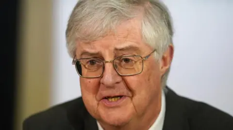 PA Media Tembakan kepala dan bahu dari Mark Drakeford mengenakan jas hitam dan kemeja putih di depan latar belakang putih.