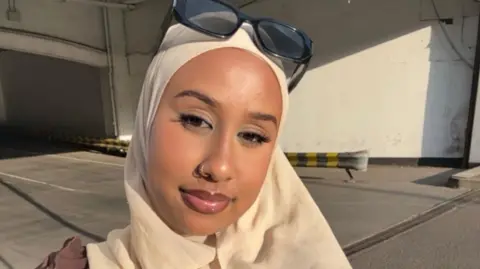 A young woman wearing a beige hijab and black sunglasses resting on her head poses for a close-up selfie outdoors 