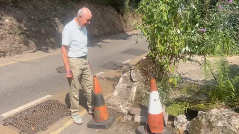Anthony Allchurch is wearing a short sleeved white shirt and observes damage to his driveway caused by a passing car.