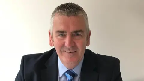 David McWilliams pictured sat against a white wall. He is wearing a blue shirt and blue tie.