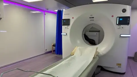 CT scanner ready to use inside a mobile unit in Coventry. The room is lit in purple and has a changing area behind a curtain in the corner.
