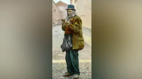 University of Cumbria A painting of Jimmy Dyer. He is standing in a cobbled street playing the violin. He is wearing a bowler hat, green coat and green trousers, carrying a satchel in front on his body.