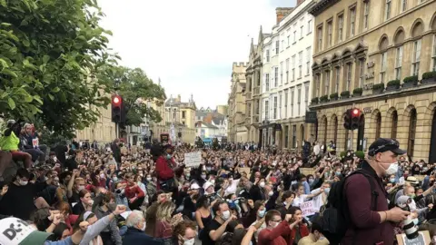 A large crowd of people, many wearing face masks and holding placards