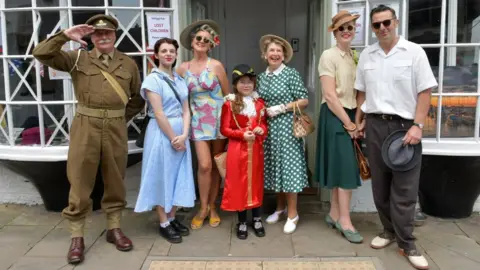 Seven people are posing for a photo dressed in mainly 1940's themed fashion. The women have styled their hair in 40's fashions and applied red lipstick. A man salutes in a khaki uniform and sports an upturned silver moustache.