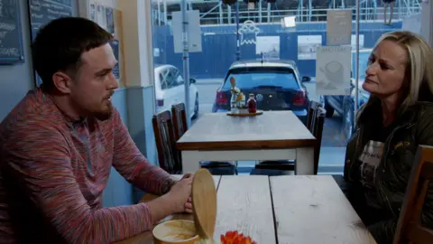 UKFS Cheri Curran, sitting in a cafe, talking to a man. She is looking at him and looks concerned. She is wearing a dark T-shirt and coat. The man has short dark hair and has on a multi-coloured top. A coffee cup is by his side. Cars are parked outside and there is building work going on outside. 