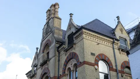 Getty Images The roof and upper storeys of Cambridge Corn Exchange, backed by a blue sky. It is a Victorian building of light-coloured brick with ornate chimneys and red and black arches over the windows.