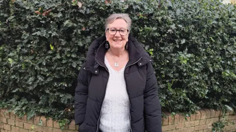 Jane Jackson is standing in front of a hedge outside, smiling to camera,  wearing a black puffer coat and a white jumper with a silver necklace