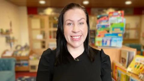 Jordan Coates is smiling at the camera with a classroom in the background. She has long brown hair and is wearing a black top with puffed shoulders.