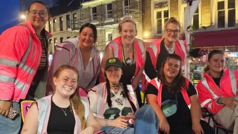 A group of eight women of varying age are gathered in Newcastle's Brigg Market at night. All of them are wearing pink high-visibility jackets. Some are stood whilst others sit in folding chairs.