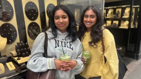 Two girls wearing grey and yellow hoodies stand in front of a jewellery shop 