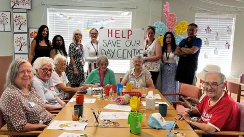 Group of older people sitting, doing art and crafts on the table and the staff is behind it and showing the sign 'Help Save Our Daycare'