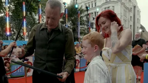 Tom Hanks is standing on a red carpet and holding and looking at the framed winning design. Tommy is also looking down at the painting and smiling. Crowds of people can be seen around them, and the edge of some Toy Story 5 branding can be seen behind Tommy and a woman with red hair who is standing next to him and smiling.