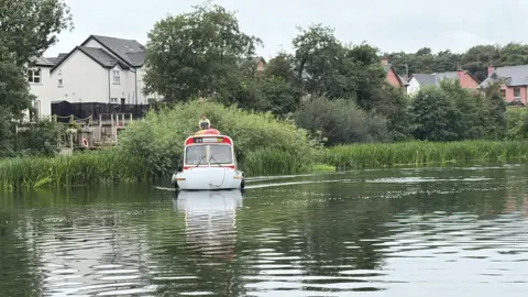 The bus boat from a distance on the River Bann sailing along. Behind it are long reeds and behind them are trees and houses.