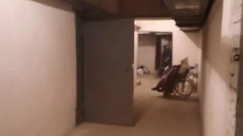 Viktoriia Izviekova Image shows the bunker Viktoriia was staying in for the first two weeks after the war started. it is slightly blurry and dark. There is a corridor which has white walls. Halfway along the corridor, there is a dark grey metal door. The door is open. Behind the door, there is a wheelchair with a person sat in it. There is an outline of another person further along the corridor too. 