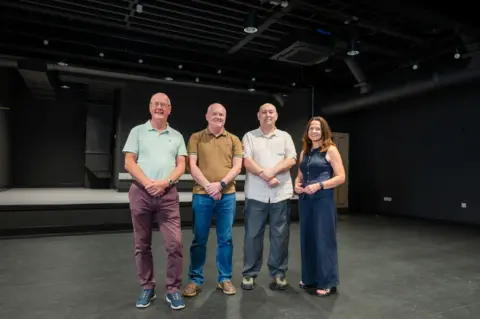 Tom Kay Photographic Catherine Eve with three men, members of Maryport Horizons, which will manage the venue. They are standing in the empty performance area inside the Carlton. The walls, ceiling and floor are completely black, with some spotlights