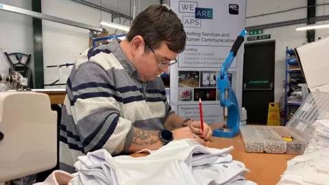 A factory worker is sitting down at a desk, busy making bibs for a baby bundle. He has short dark hair and glasses, and is wearing a striped jumper. 