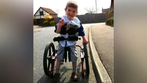 PA Media Jack Moate is standing in a frame with wheels. He is wearing an American-style baseball top, knee-length shorts and is smiling at the camera. He is next to a kerb and there is a mock-Tudor style garage in the background.
