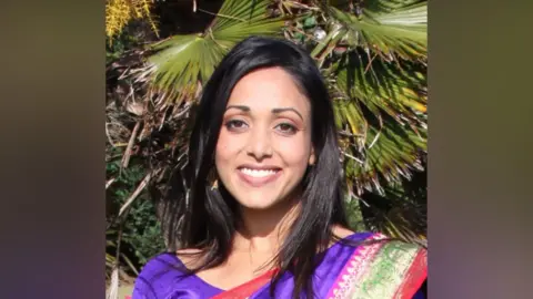 Nilu is smiling to camera in a close-up shot. She is wearing a bright purple, traditional Indian-style dress and has long black hair. There is greenery behind her