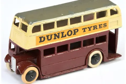 Vectis Auctions A small model of a double-decker bus. The bottom level is maroon and the top is painted cream. There are holes for windows and grey detailing to depict a metal grill on the front. A painted advert on the side reads: "Dunlop Tyres".
