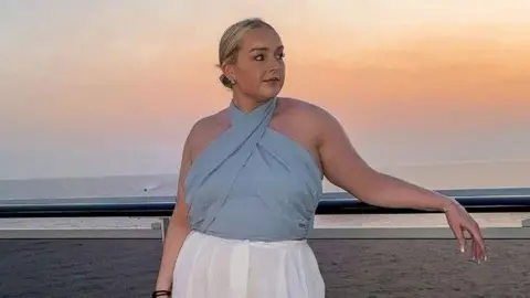 Handout Jessica Thorpe looking at the distance while leaning back on a parapet at sunset. The beach and the sea are behind her. She is wearing a blue top and white trousers and her hair is pulled back.