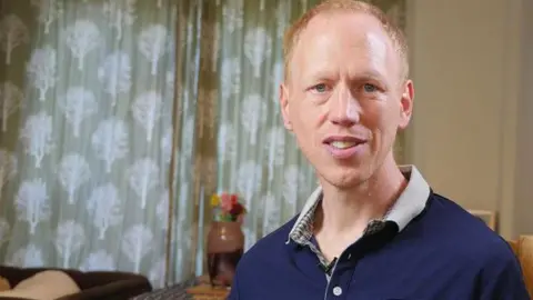 A man with short ginger hair and light blue eyes looks straight ahead. He wears a blue polo shirt with a white collar. In the background, blue patterned curtains and a vase with flowers can be seen.