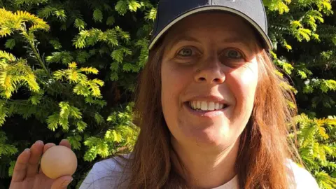 Jo Willis Jo Willis holds the egg in her hand. She is smiling at the camera as she stands outside in front of a large green bush. She wears a navy cap, black glasses, a white top and has long brown hair.